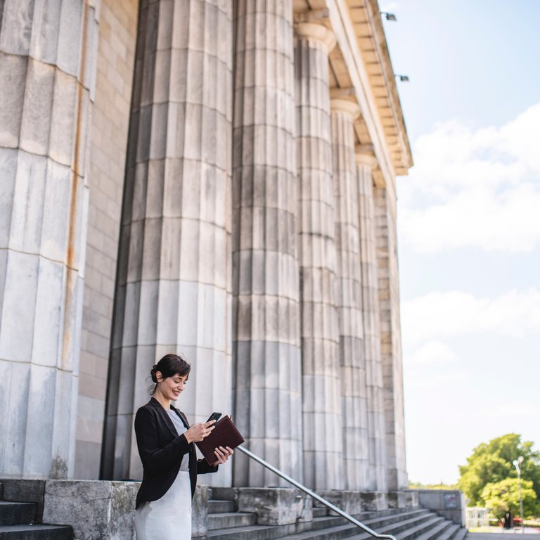 Sustaind A woman in a black blazer stands on steps, holding a tablet near large stone columns under a blue sky.