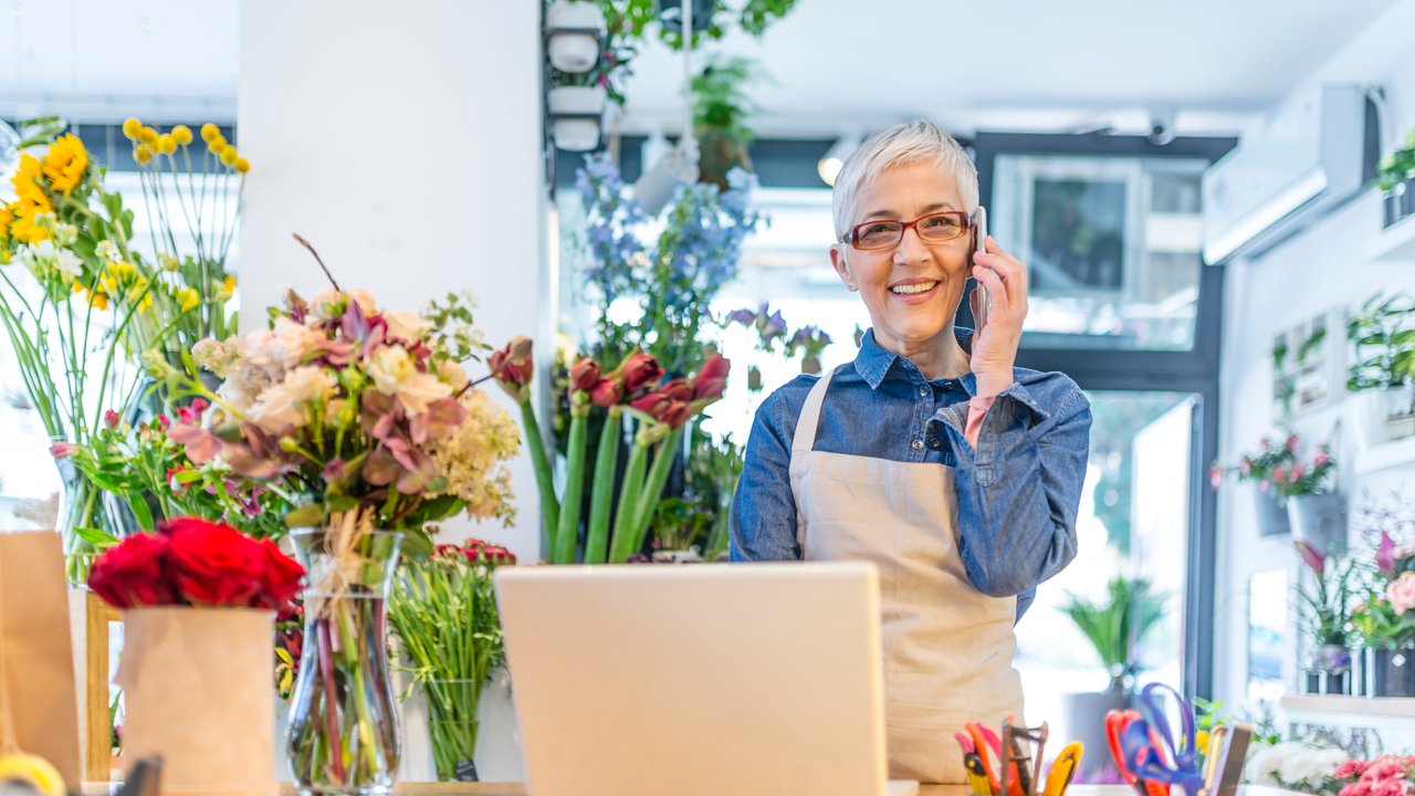 Miscellaneous Professional Liability Insurance Mature, gray hair, Flower shop worker using a smartphone and a laptop computer in her workplace and smiling while wearing denim shirt and apron. People, business, sale and floristry concept. Photo of Smiling female florist taking order on mobile phone at flower shop.